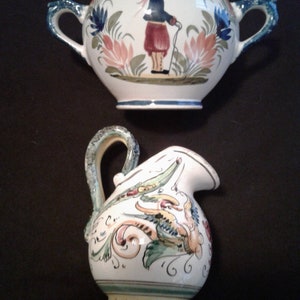May include: Two ceramic pieces, a white pitcher with a green handle and floral designs, and a white sugar bowl with a green handle and a painted figure.