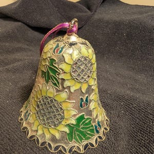 May include: A gold-colored bell ornament with a decorative design of sunflowers and leaves in shades of green, yellow, and blue. The bell has a scalloped edge and a purple ribbon.