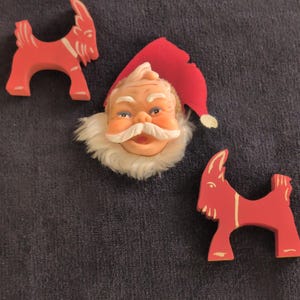 May include: Christmas decorations featuring a Santa Claus face with a red hat and two red wooden goat figurines. The Santa face has a white beard and mustache. The goats have white accents.
