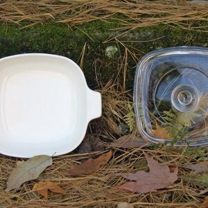 May include: A white ceramic baking dish with a glass lid. The dish is square and has rounded corners. The lid is clear and has a slightly raised rim.