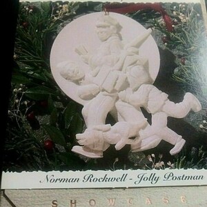 May include: A white bisque porcelain ornament depicting a scene from Norman Rockwell's "Jolly Postman" painting. The ornament features a postman delivering a package to a family on a snowy day. The ornament is part of the "Portraits in Bisque Collection" and is displayed in a white box with the words "Showcase" printed on the front.