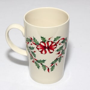 May include: A white ceramic mug with a green holly and red ribbon design. The mug has a handle and is perfect for hot beverages.