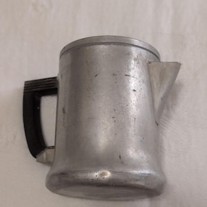 May include: A vintage aluminum coffee pot with a black handle and a spout. The pot has a cylindrical body with a lid and a handle on the side. The surface of the pot shows signs of wear and use.