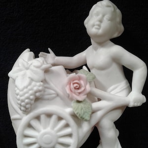 May include: A white porcelain figurine of a cherub pushing a wheelbarrow with a pink rose and green leaves. The wheelbarrow is filled with grapes.