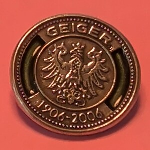May include: A round metallic pin featuring a central embossed eagle emblem. The text GEIGER is arched across the top, and the years 1906-2006 are inscribed along the bottom edge. The design includes a beaded border and a dark recessed background.