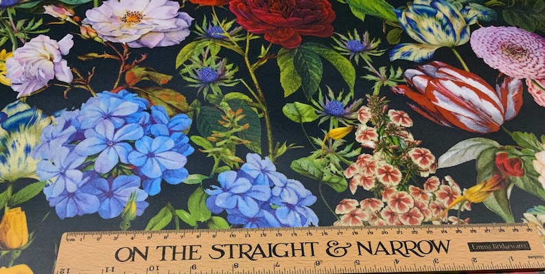 May include: A fabric with a dark background, featuring a vibrant floral pattern. The design includes roses, tulips, and other flowers in shades of red, blue, white, and yellow. A wooden ruler with the text "ON THE STRAIGHT & NARROW" is at the bottom.