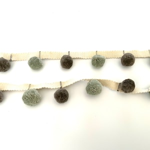May include: A white trim with a row of grey and green pom-poms. The trim is perfect for adding a touch of texture and whimsy to any project.