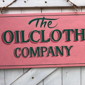 Op de afbeelding: Een roze houten bord met een zwarte rand en groene letters met de tekst "The Oilcloth Company".