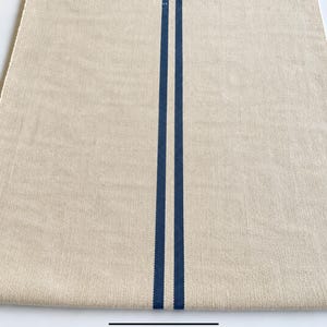 May include: A rectangular table runner made of a light beige, woven fabric featuring two parallel, vertical dark blue stripes running down the center. The textile has a visible, textured weave pattern throughout the entire surface area.