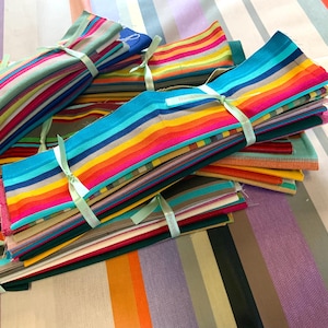 Puede incluir: Una pila de paquetes de tela a rayas de colores atados con una cinta turquesa. La tela está dispuesta en un patrón de arcoíris con tonos de rojo, naranja, amarillo, verde, azul y morado. La tela está etiquetada como "Think Outside The Box".