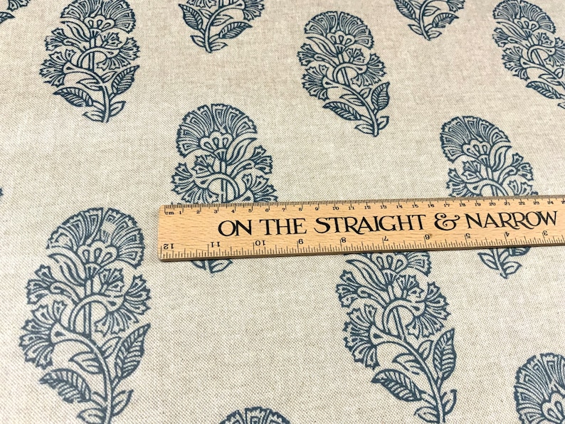 May include: A close-up of a beige fabric with a repeating pattern of dark blue floral designs. A wooden ruler with the text "ON THE STRAIGHT & NARROW" is placed on top of the fabric.