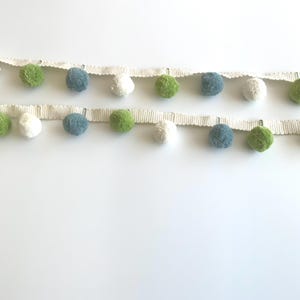 May include: Two rows of pom-pom trim with alternating colors of white, green, and blue pom-poms. The pom-poms are attached to a cream-colored, textured ribbon. This trim is suitable for various craft and sewing projects.
