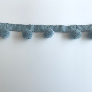 May include: A light blue trim with pom poms. The trim is made of a woven fabric and has a row of fluffy pom poms along the bottom edge.