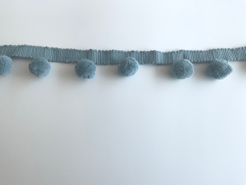 May include: A light blue trim with pom poms. The trim is made of a woven fabric and has a row of fluffy pom poms along the bottom edge.