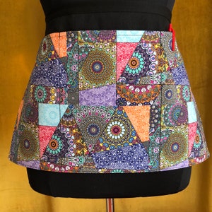 May include: A colorful patchwork apron with a geometric pattern in shades of blue, green, purple, orange, and pink. The apron has a black waistband and a red zipper.
