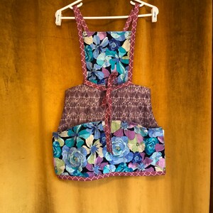 May include: A purple and blue floral print apron with a geometric pattern on the top half and a large pocket on the bottom. The apron has a tie at the waist and adjustable straps.
