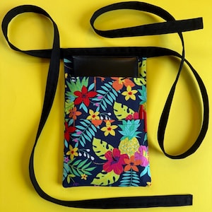 May include: A black waist apron with a colorful floral print. The apron features a dark blue background with red, pink, yellow, and green tropical flowers and leaves. A black strap extends from the top, and a black pocket is visible.