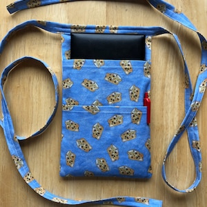 May include: A blue fabric pouch with a brown and white cookie pattern. The pouch has two pockets and a long blue strap. A black wallet and a red pen are inside the pouch.