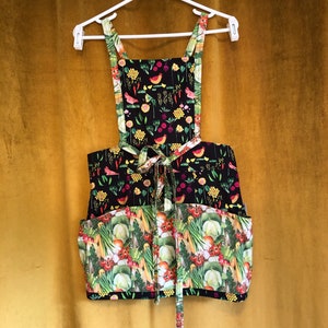 May include: A black apron with a colorful vegetable print. The apron has two pockets and adjustable straps.
