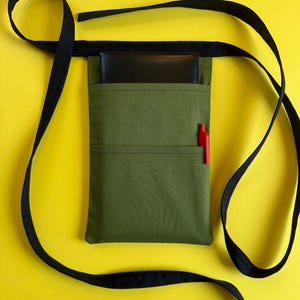 May include: An olive green server apron with a black strap, a black notepad, and a red pen. The apron has two pockets and a black strap that goes around the waist and over the shoulder. The background is yellow.