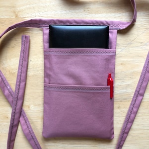 May include: A dusty rose fabric pouch with a long strap, featuring a main compartment containing a black rectangular case and a smaller front pocket holding a red pen. The edges are finished with dark zigzag stitching throughout the design.