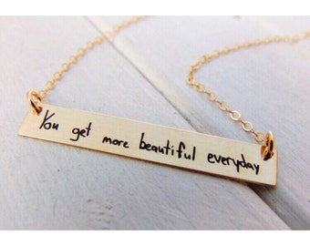 May include: Gold necklace with a bar pendant that says "You get more beautiful everyday" in handwritten font.