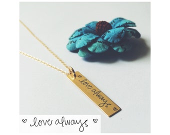 May include: Gold necklace with a long, rectangular pendant that says "love always" in cursive handwriting. The pendant is attached to a delicate gold chain.