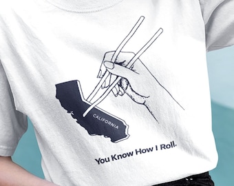 May include: White t-shirt featuring a black graphic of a hand using chopsticks to pick up a California outline. The text "You Know How I Roll" is below the graphic. The shirt is worn by a person.