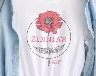 May include: White t-shirt with a pink flower graphic and the text "Zinnia's Garden Club".
