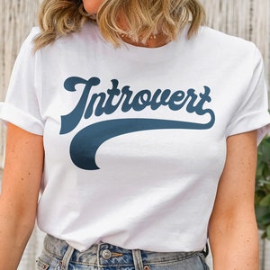Puede incluir: Camiseta blanca con un gráfico azul "Introvertido" en una fuente de béisbol retro.