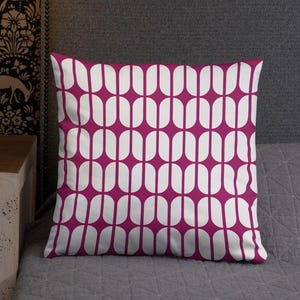 以下が含まれることがあります： マゼンタ色の背景に白い楕円が繰り返される模様の正方形の装飾用クッション。 クッションは灰色の表面に置かれ、鮮やかなコントラストを生み出しています。 デザインは幾何学的でモダンで、現代的なインテリアに最適です。