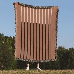 Könnte beinhalten: Eine rechteckige braune Decke mit vertikalen schwarzen und weißen Streifen. Die Decke hat einen Fransenrand und wird vor einem blauen Himmel gehalten. Die Decke wird im Freien präsentiert.