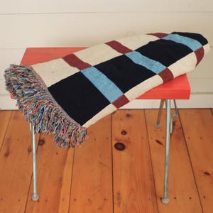 May include: A folded, rectangular throw blanket with a checkered pattern in navy, cream, burgundy, and light blue. The blanket has a fringed edge with multiple colors and is resting on a small, red stool with silver legs.