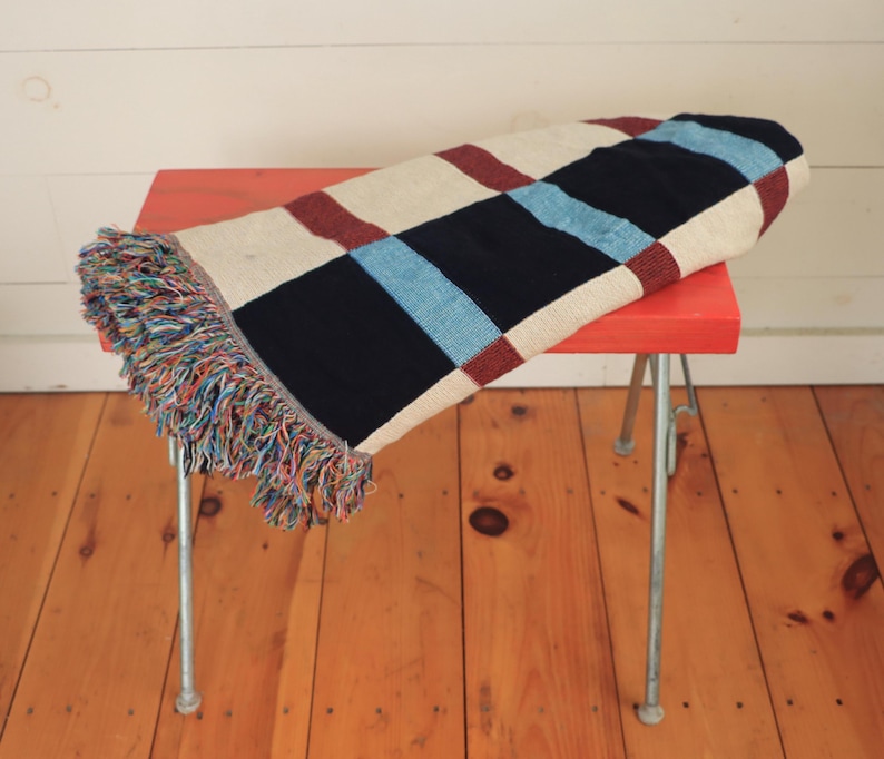 May include: A folded, rectangular throw blanket with a checkered pattern in navy, cream, burgundy, and light blue. The blanket has a fringed edge with multiple colors and is resting on a small, red stool with silver legs.
