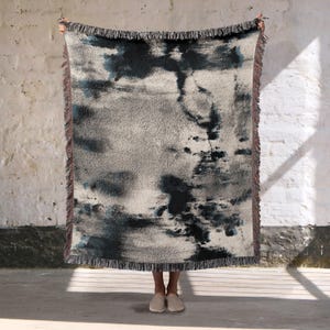 May include: A rectangular woven throw blanket with a gray and blue abstract design. The blanket has a fringed edge and is held up by a person. The color palette includes shades of gray, blue, and beige.