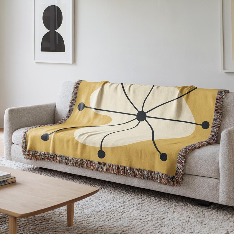 May include: Mustard yellow throw blanket with a mid-century modern design. The blanket features a cream-colored abstract shape with black lines and dots. The edges have a fringed trim. The blanket is draped over a gray sofa.