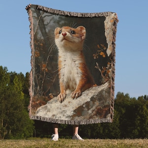 May include: A woven throw blanket with a detailed illustration of a weasel looking upwards. The weasel has reddish-brown fur and a white underbelly. The background depicts a forest scene with a blue sky.