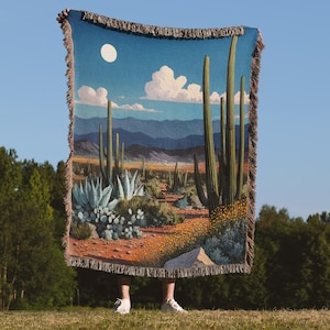 Puede incluir: Una manta de tapiz tejida con un paisaje desértico con cactus, plantas de agave y una luna llena contra un cielo azul. La manta tiene un borde con flecos y una cálida paleta de colores de naranjas, verdes y azules.
