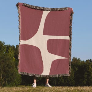 May include: A rectangular woven throw blanket featuring a deep mauve background with a large, abstract, cream-colored organic shape spanning the center. The blanket is finished with decorative fringe along the top and bottom edges.