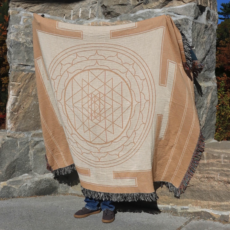 May include: Beige and brown throw blanket with a geometric design. The blanket features a large central geometric pattern, surrounded by a border. The edges have a fringe. The blanket is draped over a person.