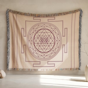 May include: Beige woven throw blanket with a geometric design in dark purple. The design features a central triangle pattern within a circle, surrounded by a square frame. The blanket has fringed edges.