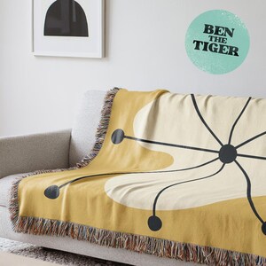May include: Mustard yellow and cream throw blanket with a mid-century modern design. The blanket features a large, stylized floral pattern in black and cream, with black circular accents. The fringe is a mix of brown, tan, and cream colors. The text "BEN THE TIGER" is on the wall.
