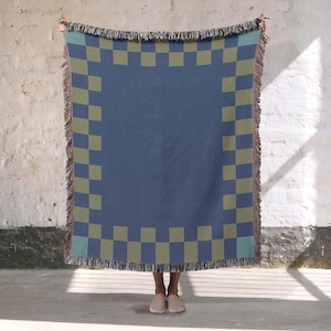 May include: A blue throw blanket with a checkerboard pattern in olive green. The blanket has a fringed edge and is held up against a white wall. The blanket is a square shape.