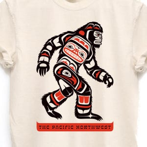 Puede incluir: Una camiseta blanca con un gr&aacute;fico negro y rojo de un Sasquatch estilizado en un estilo de arte ind&iacute;gena de la costa noroeste. El texto "THE PACIFIC NORTHWEST" est&aacute; impreso en rojo debajo del gr&aacute;fico.