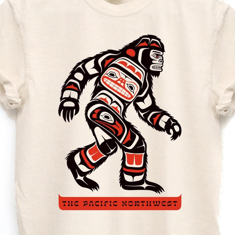 Puede incluir: Una camiseta blanca con un gr&aacute;fico negro y rojo de un Sasquatch estilizado en un estilo de arte ind&iacute;gena de la costa noroeste. El texto "THE PACIFIC NORTHWEST" est&aacute; impreso en rojo debajo del gr&aacute;fico.