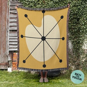 May include: Mustard yellow throw blanket with a cream-colored, abstract design featuring black lines and dots. The blanket has a fringed edge and is displayed outdoors. The design is reminiscent of mid-century modern art.