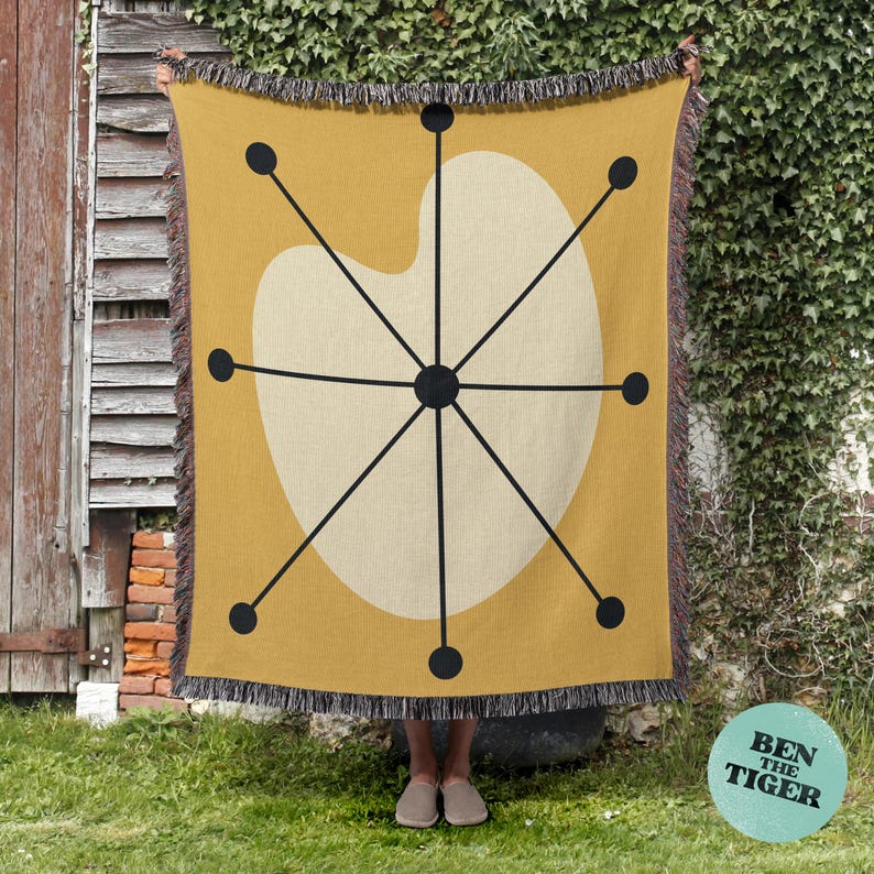 May include: Mustard yellow throw blanket with a cream-colored, abstract design featuring black lines and dots. The blanket has a fringed edge and is displayed outdoors. The design is reminiscent of mid-century modern art.