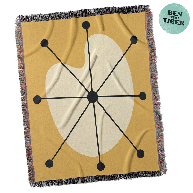 May include: Mustard yellow throw blanket with a cream-colored, abstract design featuring black lines and circles. The blanket has a fringed edge and a circular logo that reads "BEN THE TIGER".