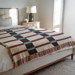 May include: A woven blanket with a plaid pattern in beige, burgundy, black, and blue. The blanket has a fringed edge and is draped over a bed. The bed has a light-colored headboard and a wooden frame.
