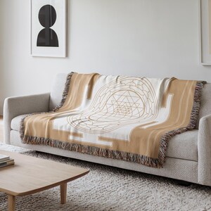 May include: A tan and white throw blanket with a geometric design draped over a gray sofa. The blanket features a detailed, symmetrical pattern in tan against a white background, with fringed edges. The sofa sits in a room with a neutral color palette.